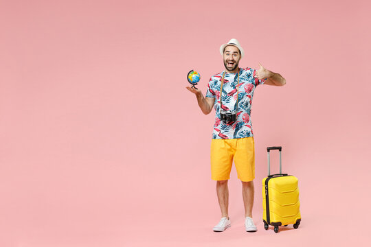 Full length of excited young traveler tourist man in summer clothes hold Earth world globe showing thumb up isolated on pink background. Passenger traveling on weekends. Air flight journey concept.