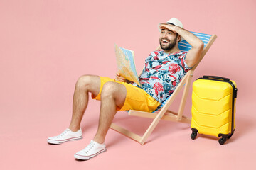 Full length of funny young traveler tourist man in hat sit on deck chair hold city map looking far away distance isolated on pink background. Passenger travel on weekend. Air flight journey concept.