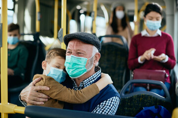 Happy senior man holding his sleepy grandson while commuting by bus during COVID-19 pandemic.