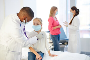 Obraz premium african male doctor checking heartbeat of kid girl using stethoscope, in hospital room. her mother is talking with assistant in the background