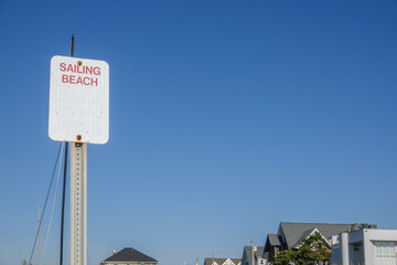 Fototapeta premium White sign on a metal post that says sailing beach against a clear cloudless blue sky.