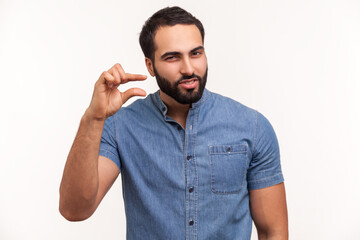 Fototapeta premium Unhappy bearded man looking disappointed and showing a little bit gesture, small amount or low scale sign, few centimeters. Indoor studio shot isolated on white background
