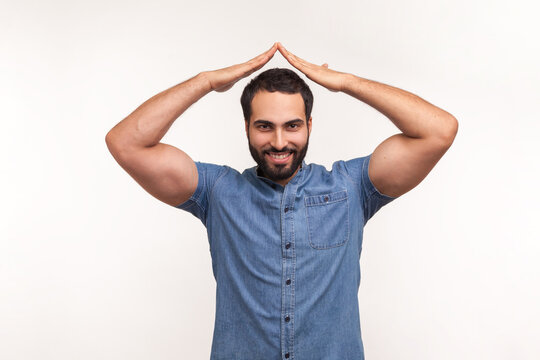 Positive Smiling Man Holding Hands Above Head Showing Roof, House Symbol, Worker Of Insurance Company Guaranteeing Safety Of Real Estate Purchase. Indoor Studio Shot Isolated On White Background