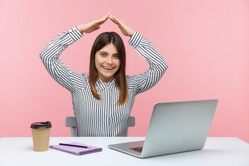 Positive smiling business woman raising hands above head showing roof sitting at workplace, freelancer, working in home office, teleworking. Indoor studio shot isolated on pink background