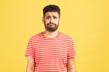 Frustrated unhappy man with beard in striped t-shirt looking at camera with sad dissatisfied expression, bad mood. Indoor studio shot isolated on yellow background