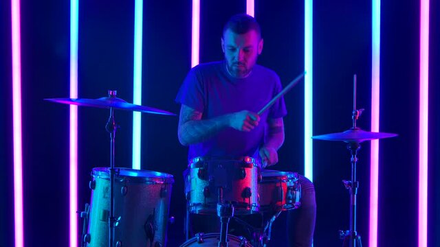 Expressive male musician plays drums in the studio against a background of multicolored neon tubes. A drummer with tattoos on his arms plays rock music with enthusiasm. Music concept. Close up.