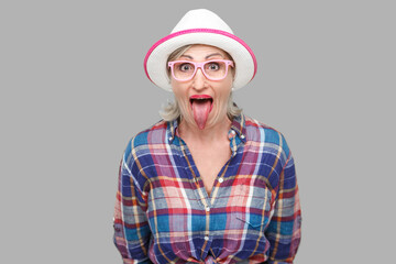 Portrait of funny surprised modern stylish mature woman in casual style with hat and eyeglasses standing tongue out, looking at camera with big eyes. indoor studio shot isolated on gray background.