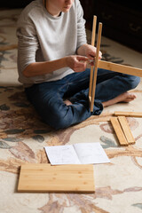 Teen boy assembles furniture according to the instructions - a wooden chair. Housework and order in it. Lesson for a teenage boy on vacation. Assistant.