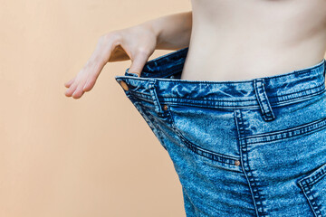 Closeup of slim waist of young woman standing in big jeans showing successful weight loss, indoor studio shot, isolated on light background, diet concept.