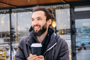 Portrait of cheerful attractive bearded young man drinking hot coffee outdoors in winter on cafe lights background