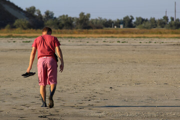 barefoot young man of the European race in a red t-shirt and pink shorts with slaps in hand goes away in the mud