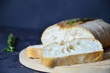Italian ciabatta bread cut in slices on wooden chopping board with herbs, garlic and olives over dark grunge backdrop, top view