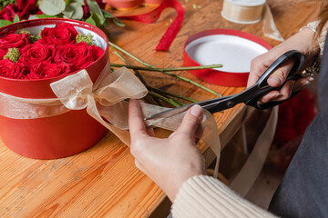 Close-up of a woman's hands cutting the ribbon from a nice surprise gift. Working with flowers on a wooden table with tools.  Entrepreneurial workspace. Red box, love gift for Valentine's Day