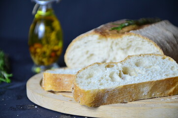 Italian ciabatta bread cut in slices on wooden chopping board with herbs, garlic and olives over dark grunge backdrop, top view