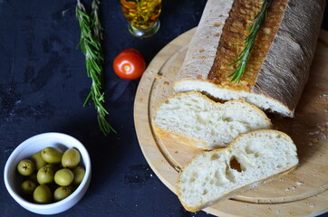 Italian ciabatta bread cut in slices on wooden chopping board with herbs, garlic and olives over dark grunge backdrop, top view