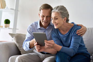 Happy old senior retired grandparents using digital tablet sitting on couch at home. Smiling mature 50s family couple enjoying websurfing online holding computer technology device making video call.