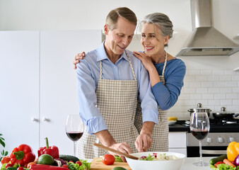Happy old middle aged 50s couple in love wearing aprons drinking wine enjoying preparing healthy diet salad nutrition at home. Smiling senior family hugging, cooking vegetable meal together in kitchen