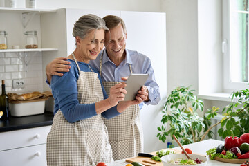 Happy middle aged 50s couple preparing salad using digital tablet in kitchen. Smiling mature older husband and wife holding computer cooking together searching healthy diet food online recipes at home