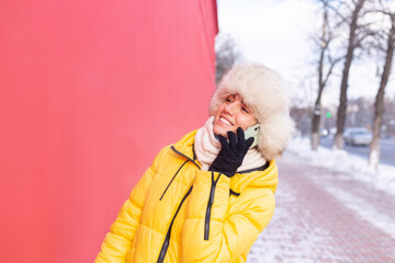 Happy young woman on a background of a red wall in warm clothes on a winter sunny day smiling and talking on the phone on a snowy city sidewalk