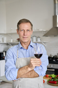 Confident Middle Aged Old Adult Single Man Holding Glass Standing In Kitchen, Vertical Closeup Portrait. Handsome Senior 60s Chef Sommelier Wearing Apron Enjoying Red Wine And Cooking At Home.