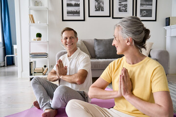 Happy healthy older senior 50s couple meditating with namaste hands at home. Smiling old middle aged husband and wife enjoying doing yoga exercises together, feeling no stress, energy, peace of mind.