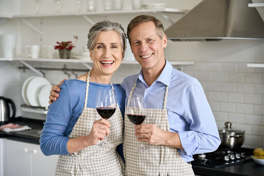 Happy old senior 50s couple wearing aprons hugging having fun drinking wine, looking at camera in kitchen. Smiling middle aged husband and wife holding glasses, cooking, celebrating together. Portrait