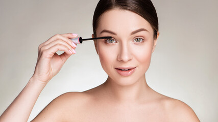 Fototapeta premium Beautiful brunette caucasian young woman prepare herself, applying mascara on her eyes with a brush. Clean, fresh, natural, flawless skin. Soft smile on her face. Close up on a neutral background.