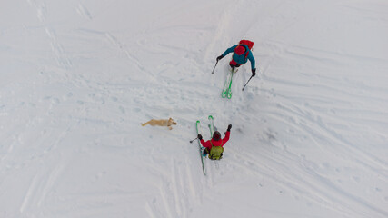 Two backcountry skiers with green skis and colorful clothing accompanied by a dog are seen from above waiting to descend into valley. Aerial drone view of skiers.