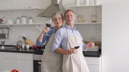 Happy affectionate older middle aged family couple wearing aprons having fun, dancing in kitchen. Senior husband and wife drinking wine enjoying dance celebrating Valentines day or anniversary at home