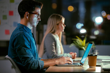 Attractive young businessman working with laptop in conference room at modern startup at night.