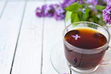 Delicate setting of the morning tea table with lilac flowers, a transparent cup of tea and a saucer and a white vase on a white wooden board. Copy space. Spring breakfast concept