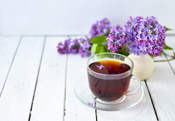 Delicate setting of the morning tea table with lilac flowers, a transparent cup of tea and a saucer and a white vase on a white wooden board. Copy space. Spring breakfast concept