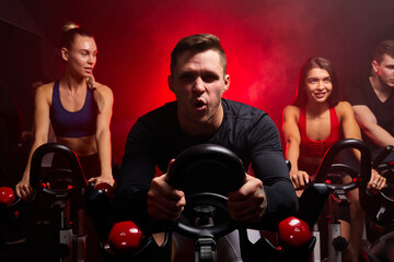 man exercising cardio training on bicycle in fitness gym with friends in the background. bodybuilder, lifestyle, exercise fitness, workout and sport training concept