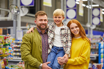 family wth son in supermarket, young parents hold cute child boy in hands, look at camera and smile, shelves with products in the background, in the aisle