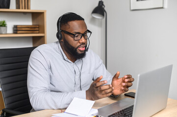 Concentered young African-American student presenting his project online on the laptop, wearing glasses and headset, making notes, focused speaker briefing the sales team on a video call