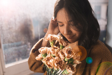 beautiful young woman smelling bouquet of pink gillyflowers at home by window. closed eyes,...