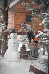 Cute boy and his little sister finishing making a snow man under the tree-house during the snowfall in winter in Russia. The put on a basket on it as a hat. Image with selective focus and toning