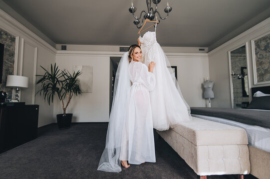A beautiful, smiling, cheerful, sexy blonde bride in lingerie, a dressing gown stands near the bed and holds a hanging white dress. Wedding portrait of a cute girl. Morning preparation.