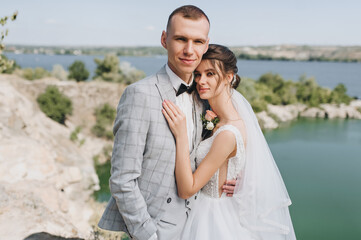 A young groom in a gray tailcoat and a beautiful curly-haired bride in a white dress are hugging on the background of a cliff, rocks and a river. Wedding portrait of newlyweds in love.