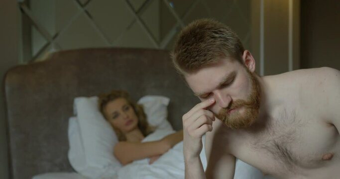 Prblems in bed. The upset ginger head man is worrying while sitting on the bed. His disappointed blonde girlfriend in thong is leaving the room.