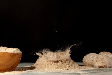 Homemade bread making with a wooden bowl of flour and a few doughs on background. Another dough is dropping into a pile of flour on marble countertop creating a flour splash against dark background.