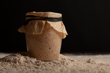 Close up side view image of glass cup of sourdough starter culture with bubbles due to active respiration. The top is covered with brown paper tightened with elastic band. The background is dark.