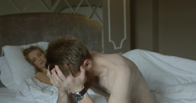 Depressed ginger head man worrying about problems in bed. The blurred background of the sad blonde woman lying on the bed and waiting.