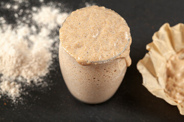 Top view of an actively rising sourdough starter culture which is overflowing over the top of the glass cup it is in. Bubbles show fermentation. The cover paper and flour is in the dark background.