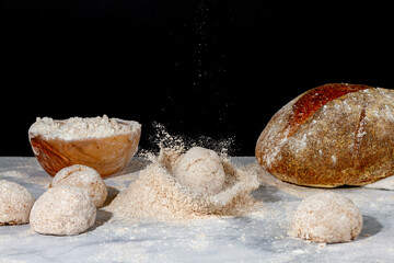 Fototapeta na wymiar A bread loaf splash on a pile of wholewheat flour on marble countertop with dark background. There is fresh sourdough bread, a wooden bowl of flour and other dough loaves on the scene. It is a mess.
