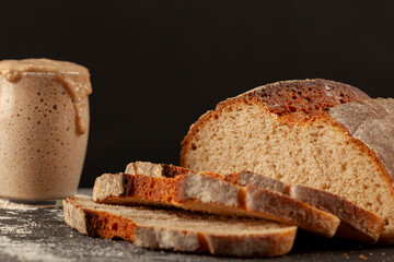 Close up side view image of a freshly baked thick crust sourdough artisan bread with an active rising sourdough starter culture in the background. Rising culture is overflowing the glass cup.