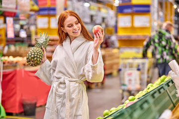 smiling female holding pineapple and apple in hands in the store, caucasian lady in bathrobe at shopping