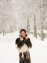 girl in a fur coat stands smiling looks up in the park, winter falling snow, walking outdoors in the park.