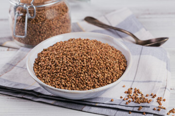 Organic uncooked scattered buckwheat grain in a bowl and glass jar on a white wooden rustic background. Healthy and diet food concept.
