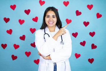 Young caucasian doctor woman wearing medical uniform and stethoscope over blue background with red hearts smiling and pointing with hand and finger to the side